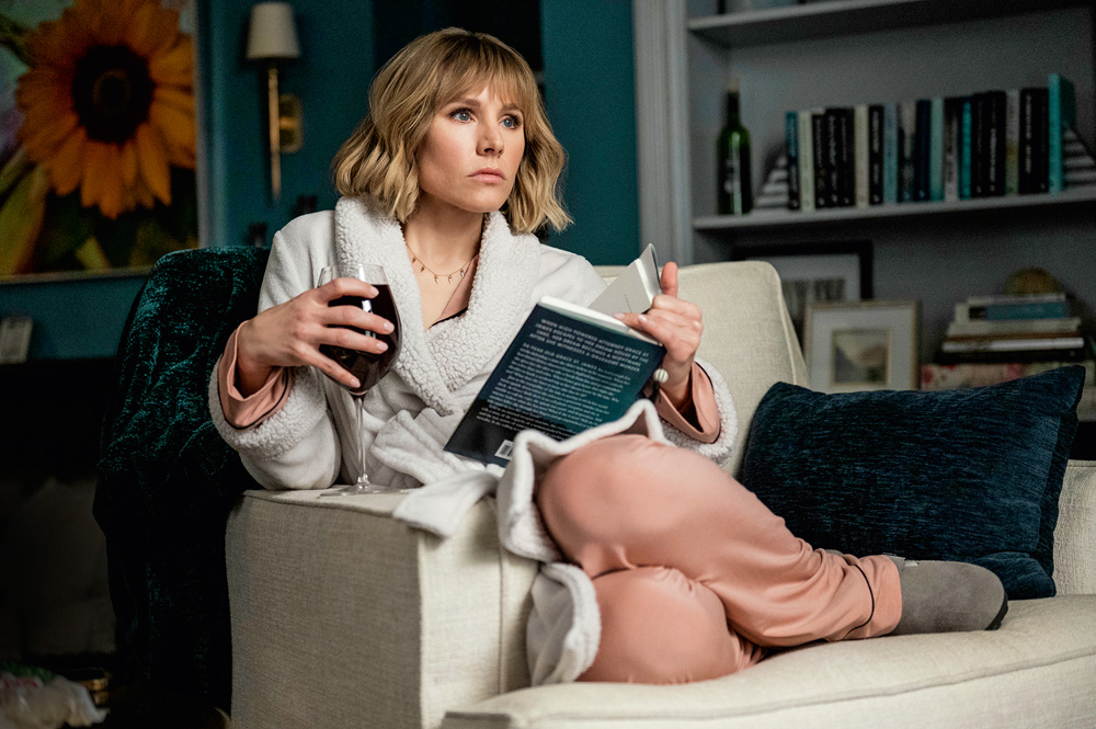Imagem mostra mulher de roupão, com um livro e uma taça de vinho na mão, sentada no sofá