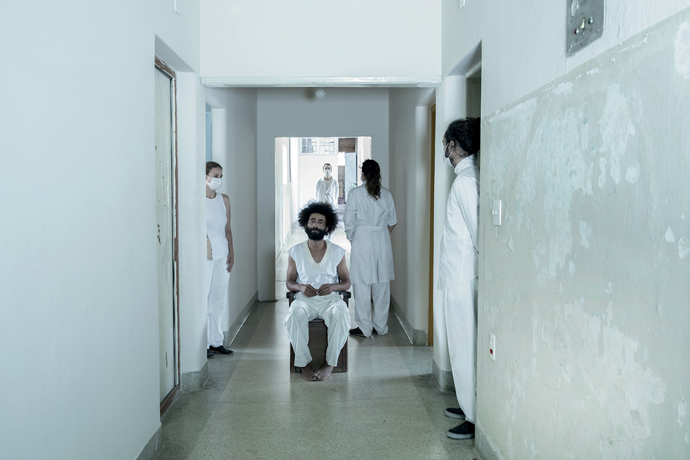 Imagem mostra corredor com homem sentado ao centro, de roupas brancas. Outras pessoas com roupas de enfermeiro estão ao seu redor.