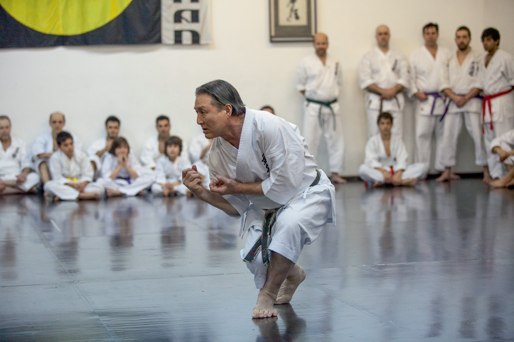 Foto atual de Taketo na academia em Pinheiros. Ele está no centro e alguns alunos o observam encostados na parede.