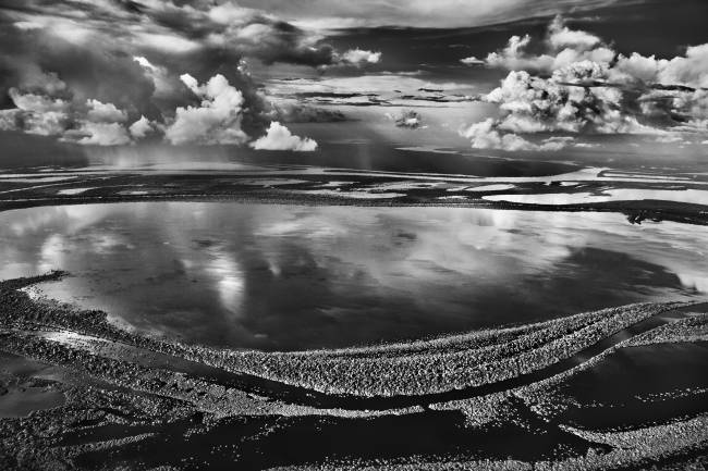 Foto em preto e branco de um rio calmo, com um dezena de nuvens no céu