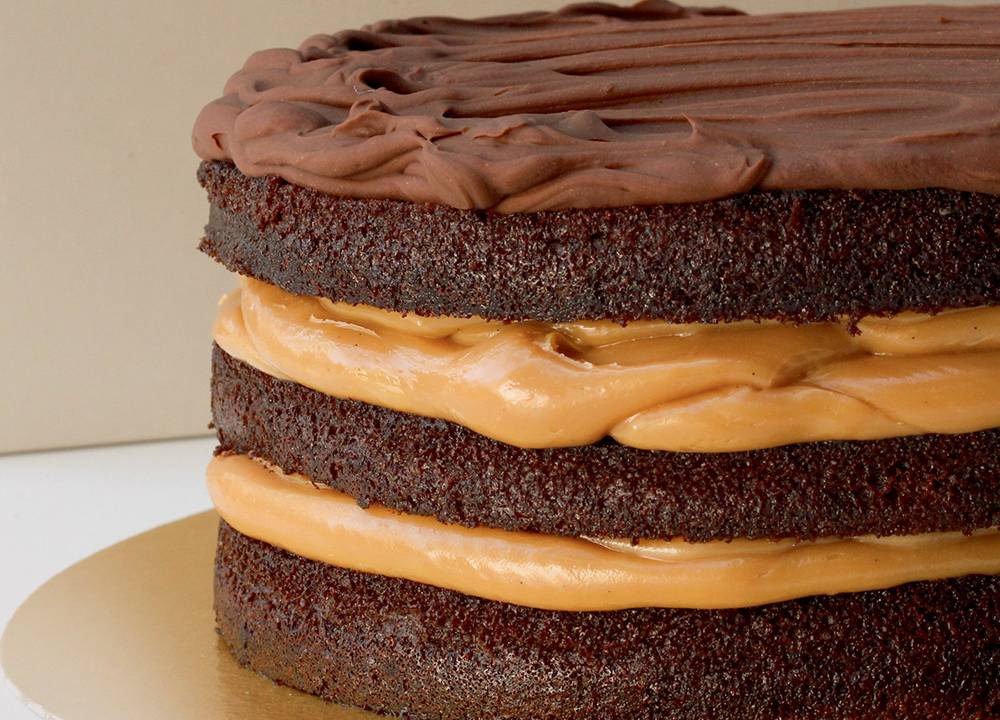 Visão lateral de um bolo dividido em camadas, recheado por doce de leite, com ganache em seu topo e massa escura