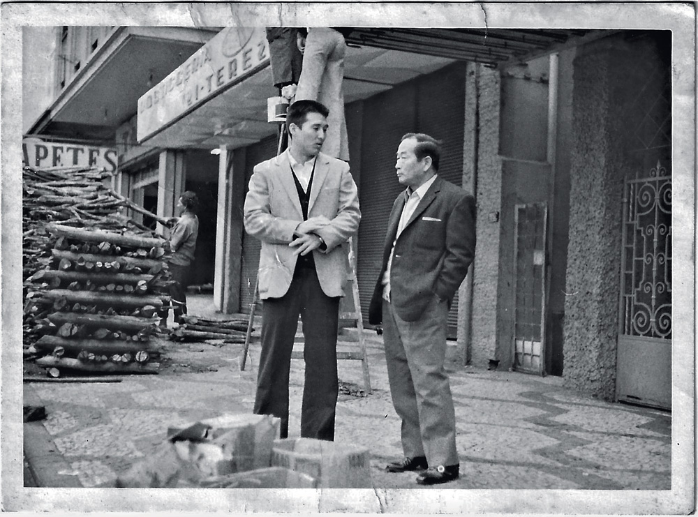 Foto em preto e branco mostra dois homens japoneses nas ruas de São Paulo. Ambos trajam um terno e conversam entre si.