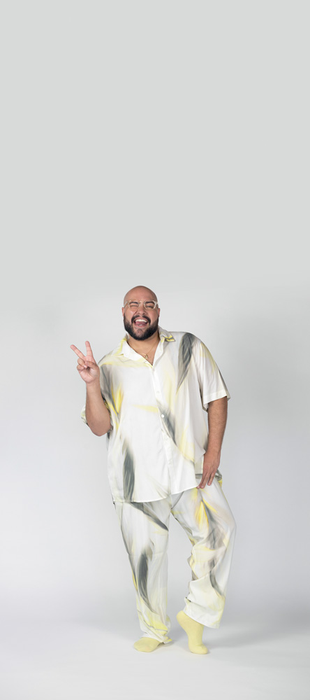 Retrato de corpo inteiro de Tiago Abravanel. Ele usa um conjunto de pijama branco com manchas amarelas e cinzas.