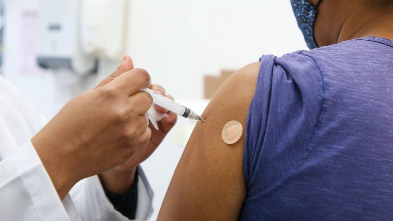 Imagem mostra braço de pessoa recebendo uma vacina. Só é possível ver as mãos da enfermeira aplicando a seringa.