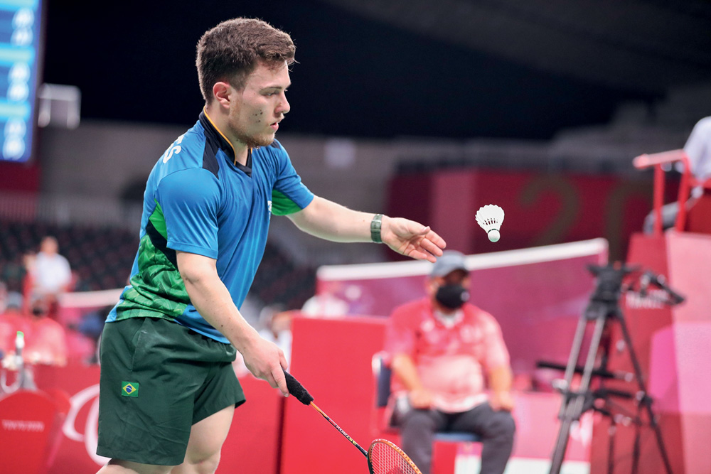 Imagem mostra homem de camiseta azul e verde com uma raquete na mão, preparando para bater em uma peteca.