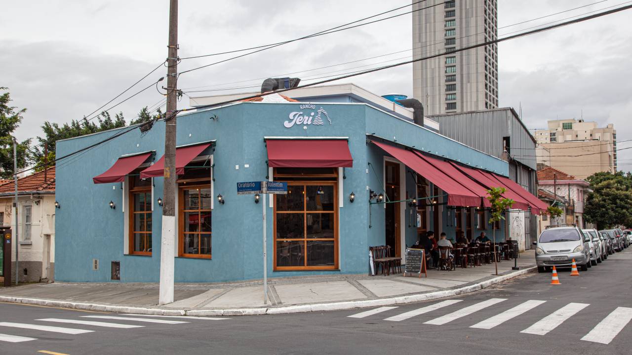 Bar de esquina com paredes pintadas em tom de azul. Há mesas na calçada e toldos de cor vermelha sobre as janelas.