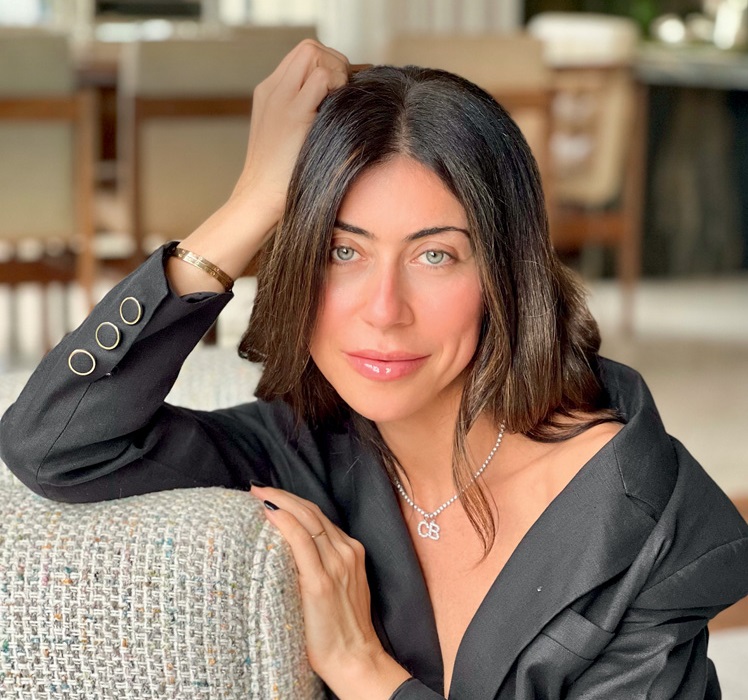 Carol Bassi posa com uma mão na cabeça e a outra apoiada no sofá. Veste roupa preta e tem cabelos longos.