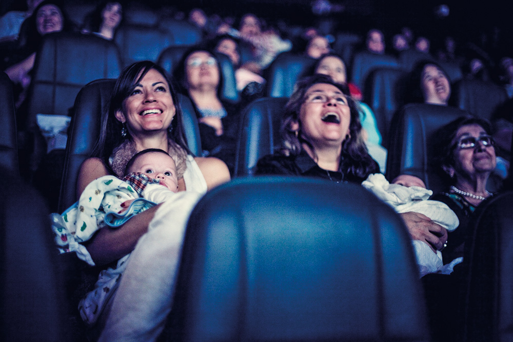 Foto mostra duas mães no cinema segurando seus bebês. Elas riem do filme.