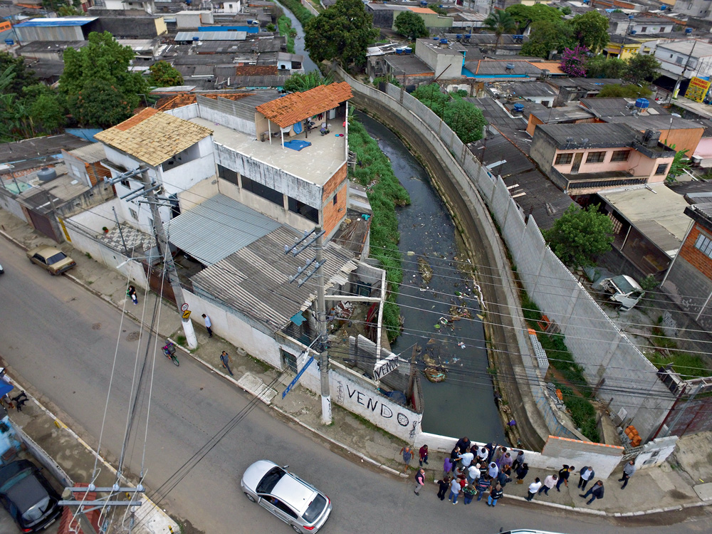 Imagem mostra rua passando sobre córrego, que passa entre diversas casas.