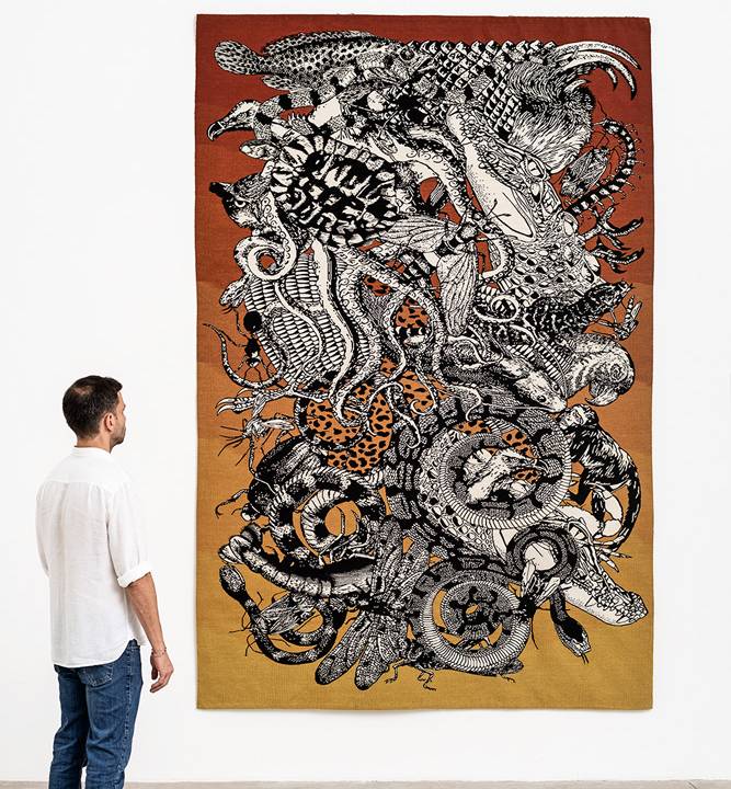 Homem observa tapeçaria em tons de preto, branco, amarelo e vermelho exposta em uma galeria. Os desenhos são de animais selvagens.