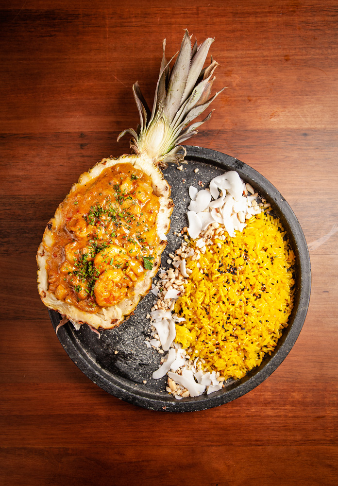 Prato raso de cor preta com abacaxi cortado ao meio e recheado por curry de camarão acompanhado de farofa de cor amarela e lascas de coco