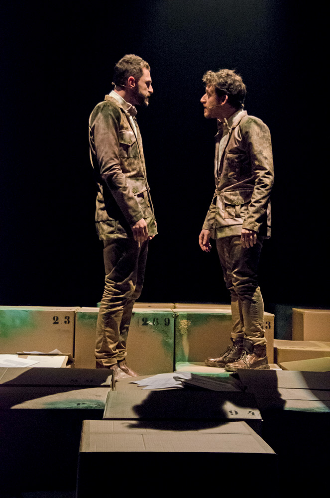 Dois atores encenam no palco com roupas surradas na cor marrom. Eles estão virados um de frente para o outro