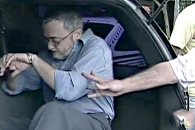 Imagem mostra homem de cabelo grisalho e camisa azul em porta-malas de carro.