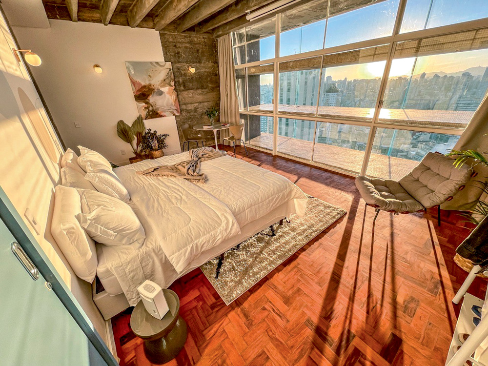 Imagem mostra quarto com cama de casal e chão de taco iluminado por pôr do sol que entra pela enorme janela que dá para uma vista de prédios de SP.