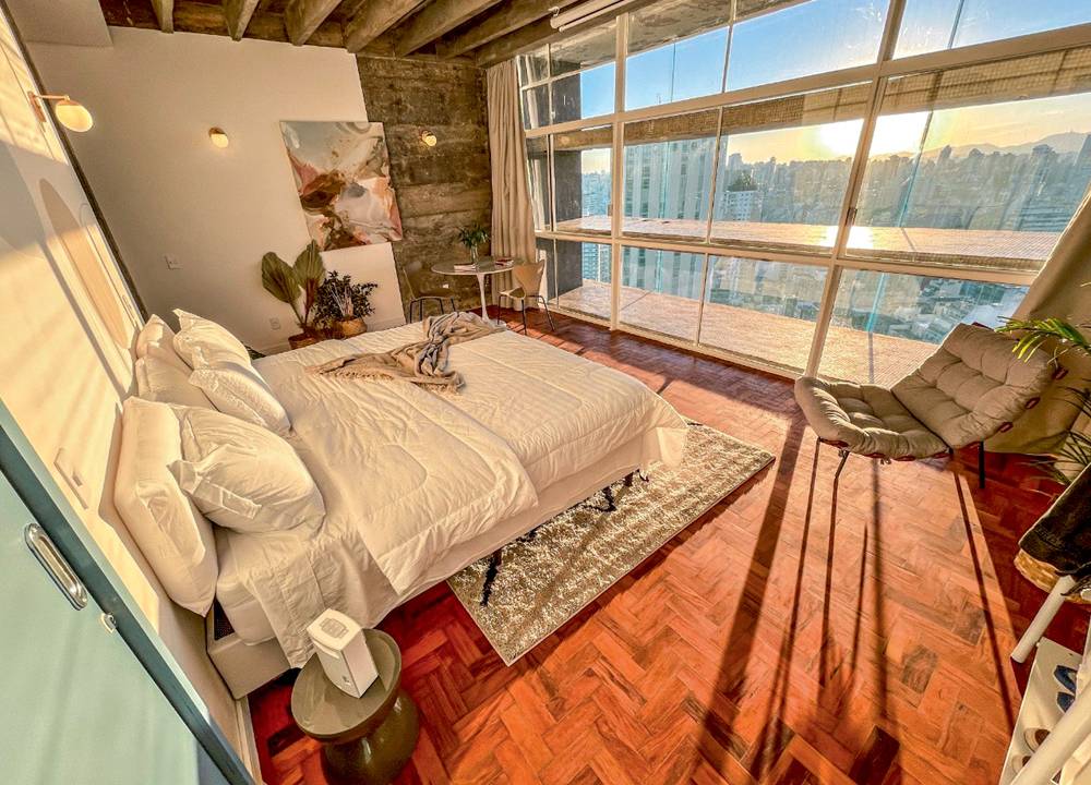 Imagem mostra quarto com cama de casal e chão de taco iluminado por pôr do sol que entra pela enorme janela que dá para uma vista de prédios de SP.