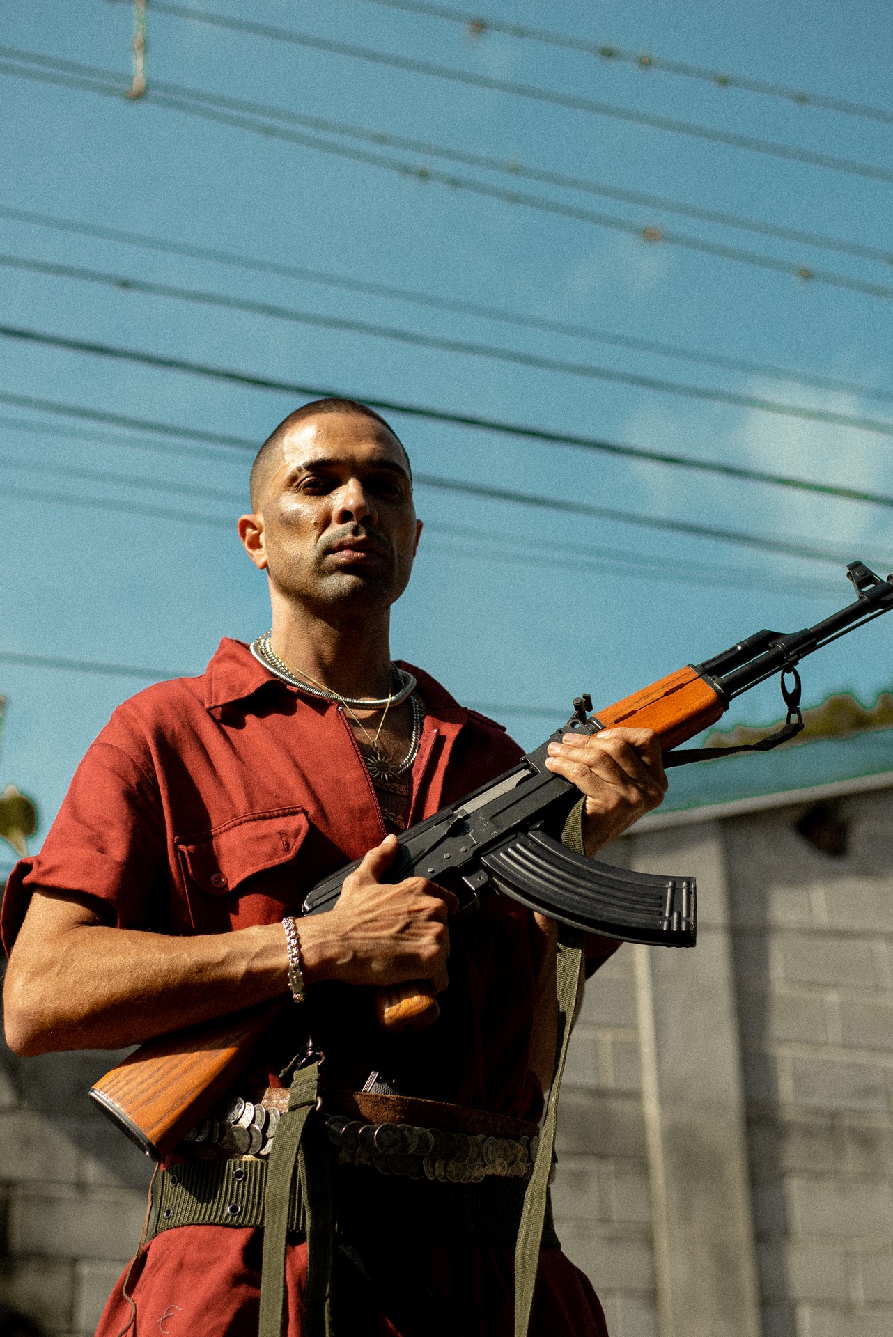 Imagem mostra homem de camisa vermelha segurando um fuzil. Ao fundo, céu azul.