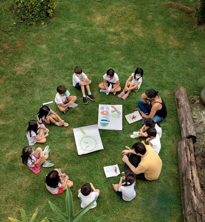 Imagem mostra círculo de crianças em gramado, com folhas de papel desenhadas ao centro.