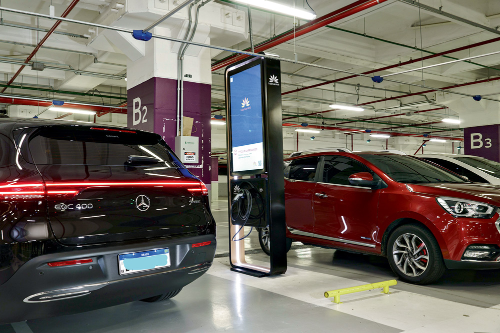 Imagem mostra dois carros, um preto e outro vermelho, sendo carregados em estação de carregamento elétrico, em estacionamento.