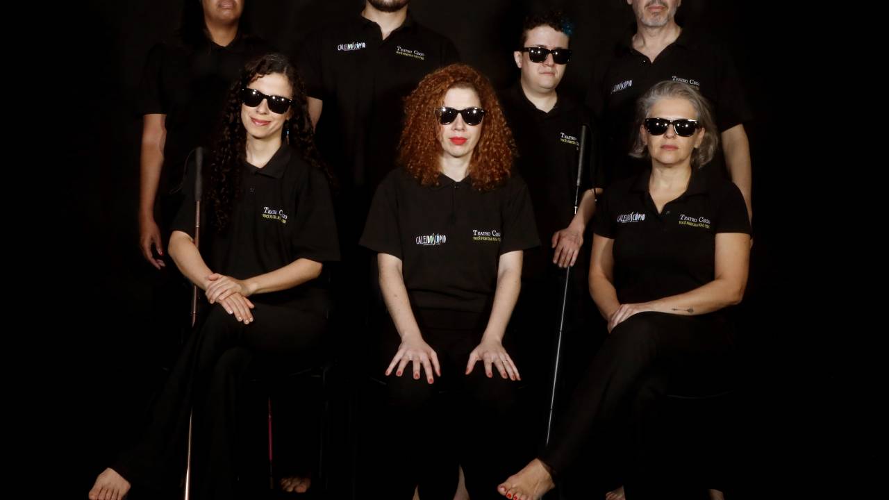 Foto mostra sete atores, todos vestidos de preto e com óculos escuros. Três deles possuem deficiência visual. Quatro, uma mulher e um homem, estão em pé atrás. Três, todas mulheres, sentam-se em cadeiras na frente. O fundo é preto.