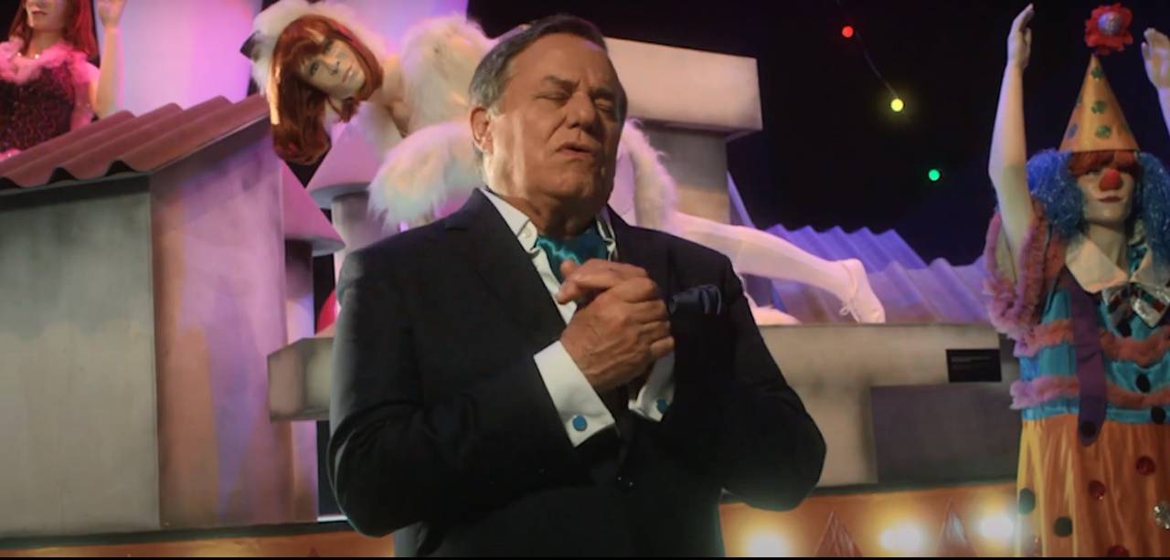 Ronnie Von em cena de clipe produzido com Rita Lee veste terno, segura as mãos juntas de olhos fechados.