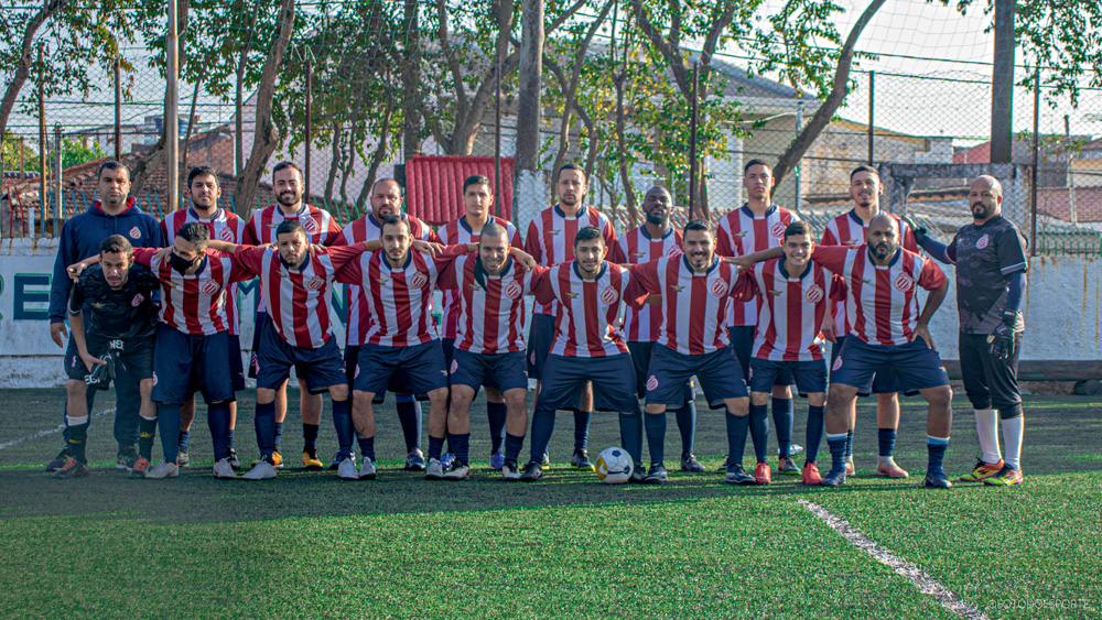 Imagem mostra time completo de futebol, com uniforme listrado vermelho e branco, enfileirados e abraçados.