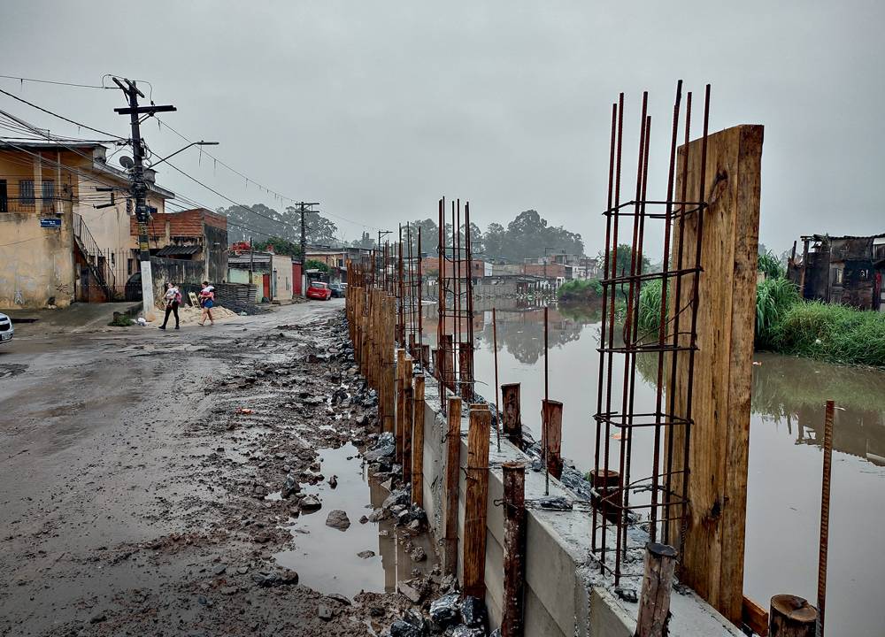 Imagem mostra córrego ao lado de rua de terra, esburacada e repleta de poças.