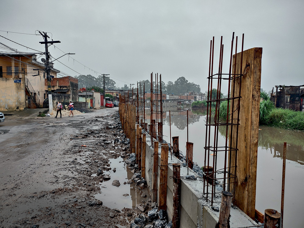 Imagem mostra córrego ao lado de rua de terra, esburacada e repleta de poças.