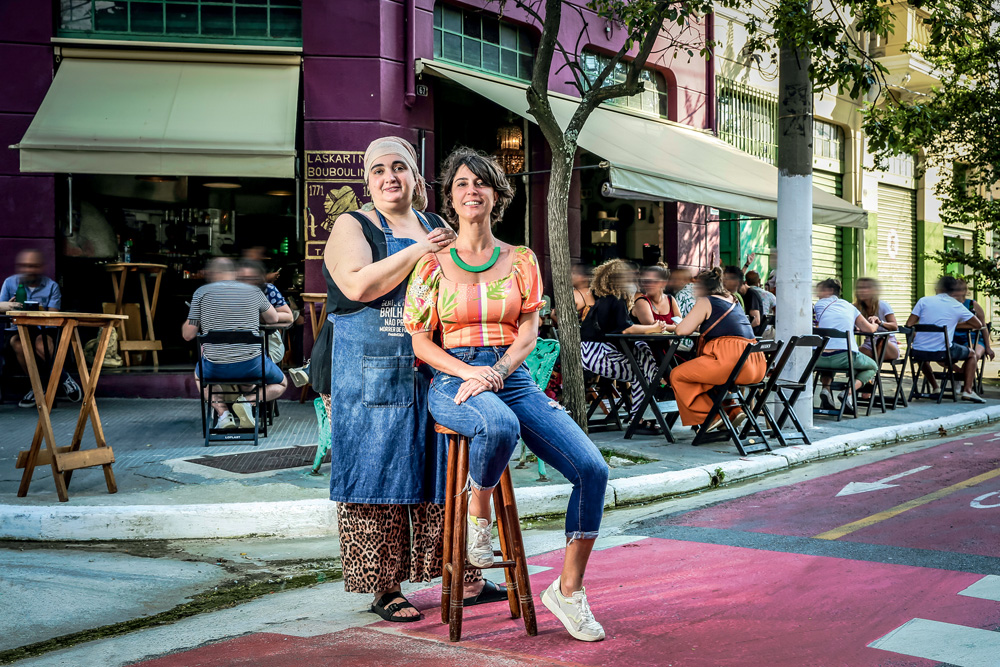 Duas mulheres, donas do empreendimento, posam na frente de um bar na Barra Funda. O local está movimentado. Ambas são brancas e usam jeans.