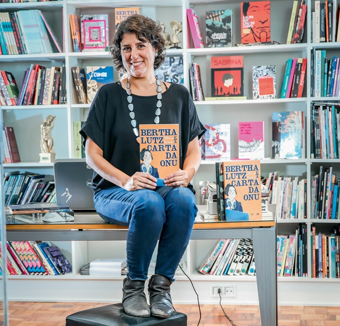 Leticia de Castro. da Editora Veneta, posa sentada segurando livro de capa laranja. veste jeans e camiseta preta. Ao fundo, estantes de livro.