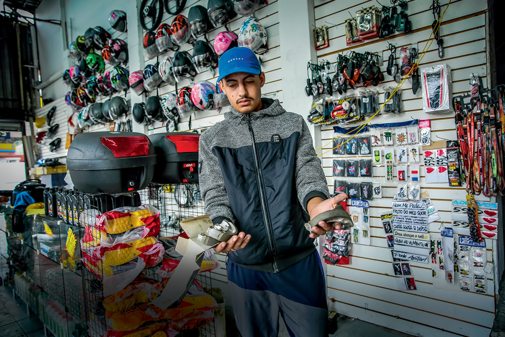 Imagem mostra homem de boné azul e roupa cinza com as mãos abertas, segurando produtos. Ao fundo, parede com diversos capacetes de moto à venda.
