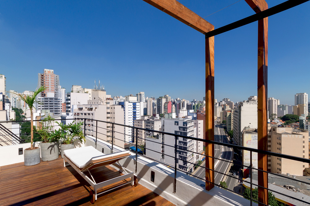 Imagem mostra terraço com chão de madeira e cadeira de praia. Um horizonte de prédios sob um céu azul preenche a imagem.