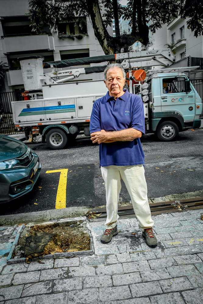 Imagem mostra homem grisalho de camisa polo azul e calça branca com os braços cruzados, ao lado de raiz de árvore que caiu.