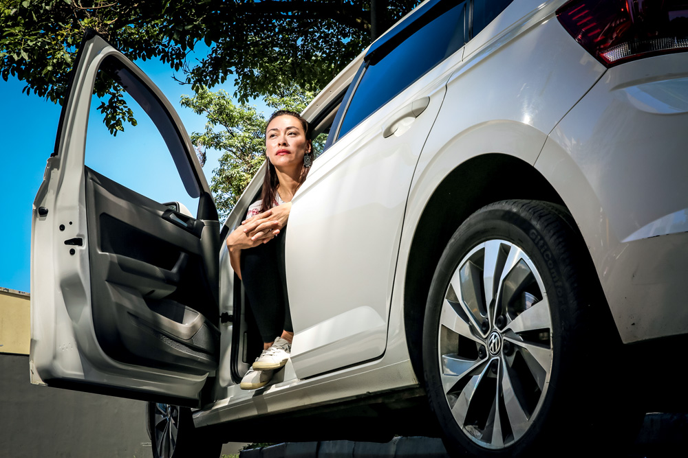 Imagem mostra mulher com os braços encolhidos sobre as pernas, no banco do motorista de um carro cinza. A porta está aberta.