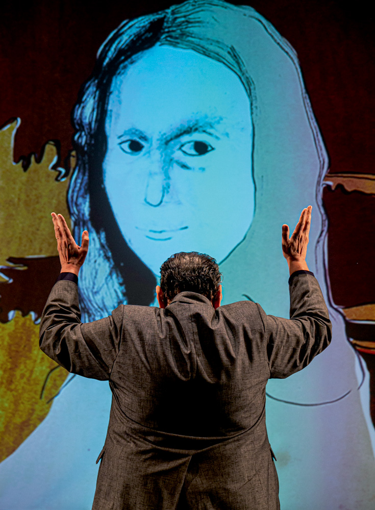 Ator Cacá de Carvalho aparece de costas, levantando os braços e fazendo gestos com as mãos, em frente a uma projeção de Mona Lisa.