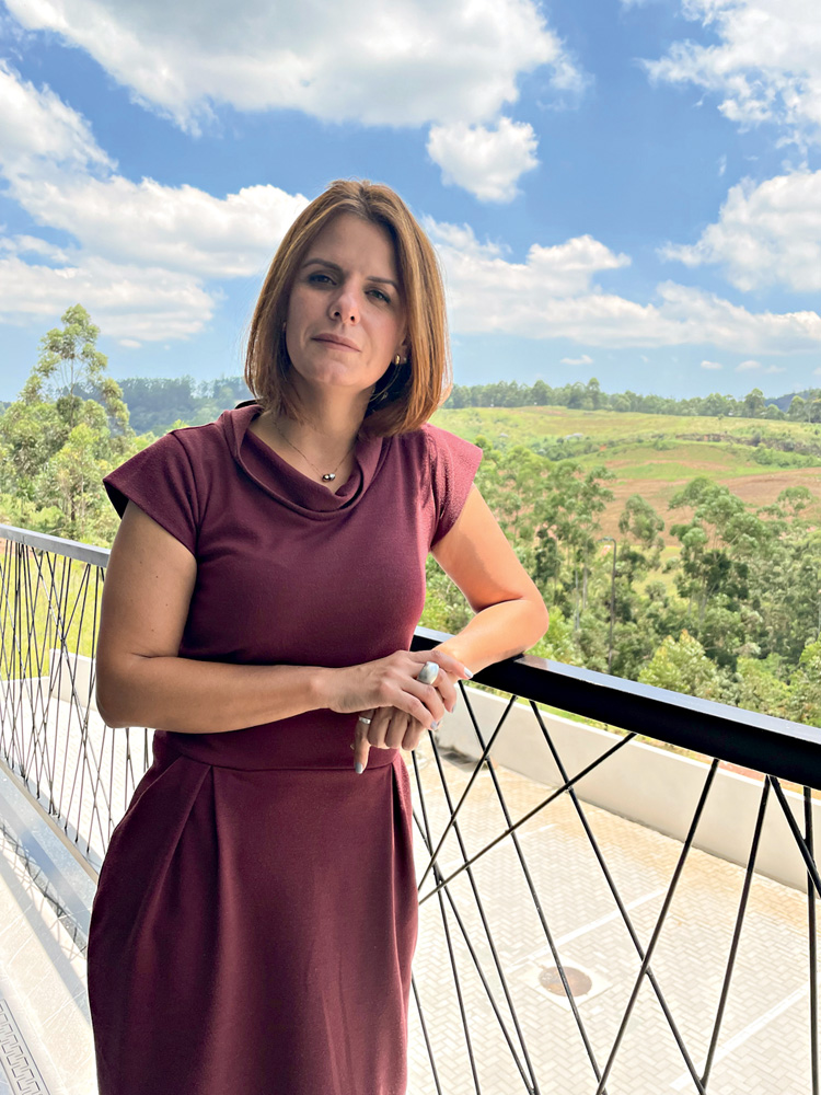 Adriane posa em uma sacada. Ao lado é possível ver um campo verde. Ela é uma mulher branca, morena e usa um vestido vermelho.