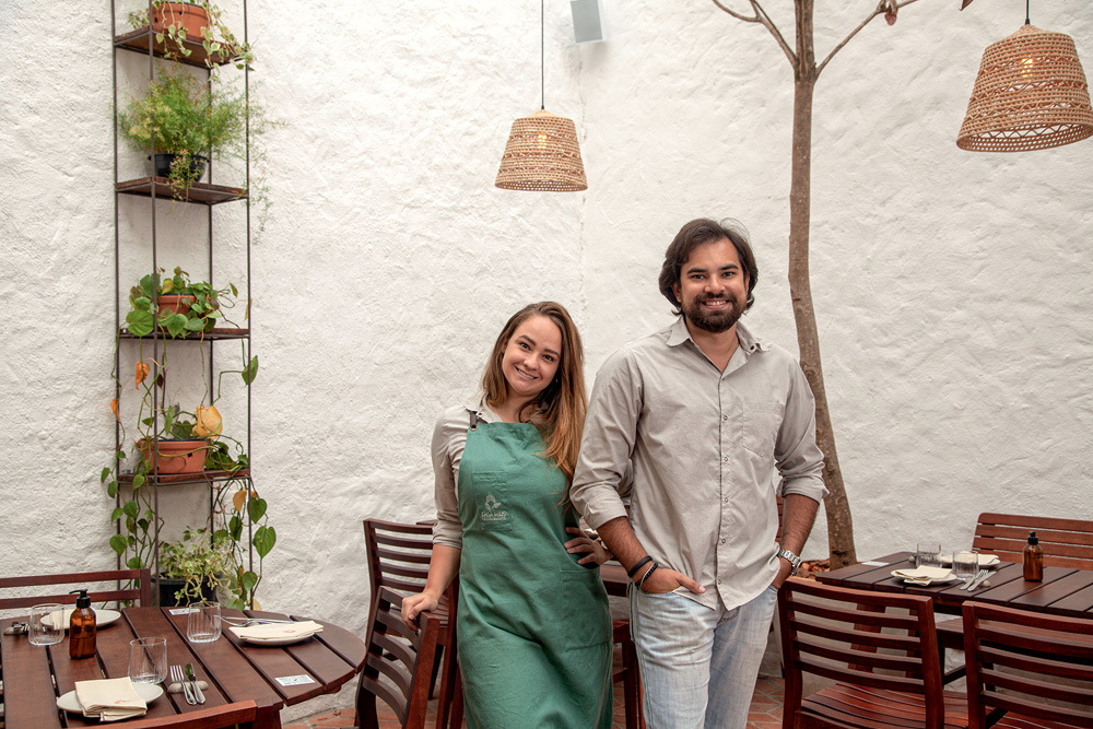 Chefs Rodrigo Aguiar e Giovanna Perrone posam lado a lado entre mesas de madeiras em frente a uma parede de cor bege