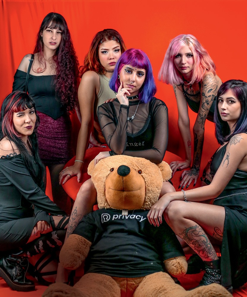 Seis mulheres vestem preto e posam ao lado de um urso de pelúcia com a camiseta da marca. Algumas têm cabelo colorido, piercings e tatuagens.