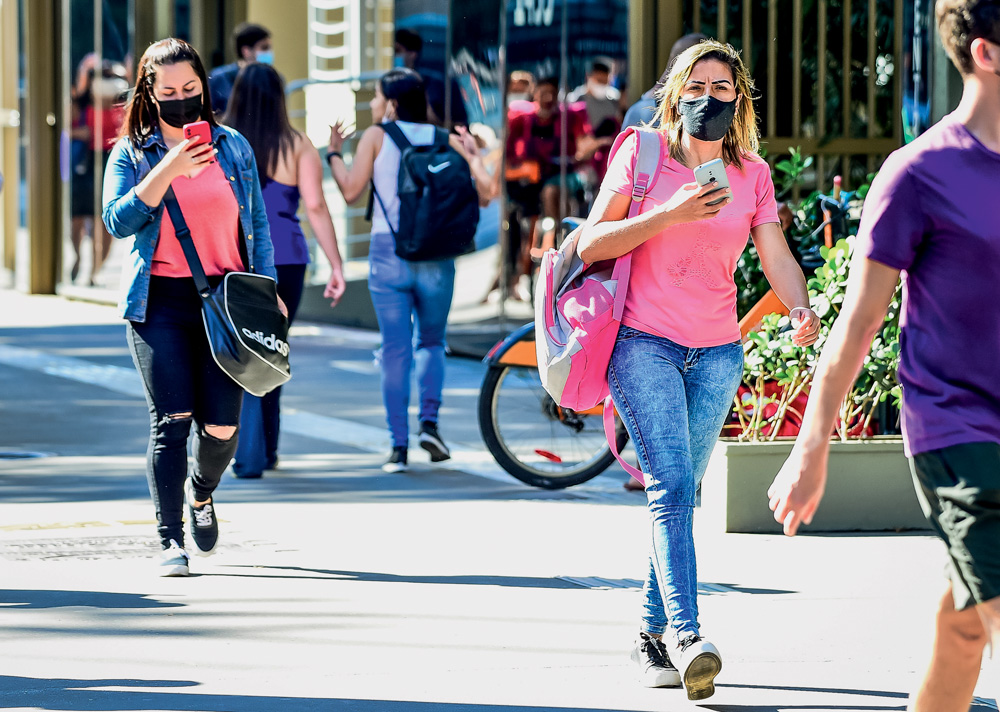 Imagem mostra pessoas andando na calçada com os celulares nas mãos.