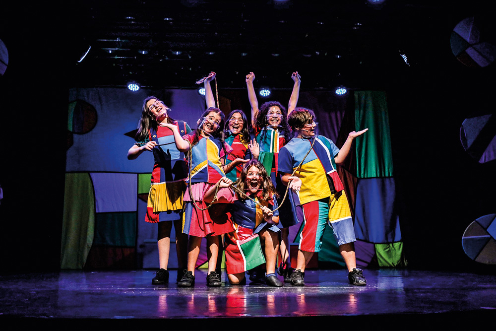 Seis crianças, cinco meninas e um menino, posam no palco. Eles usam figurinos coloridos e máscaras transparentes.