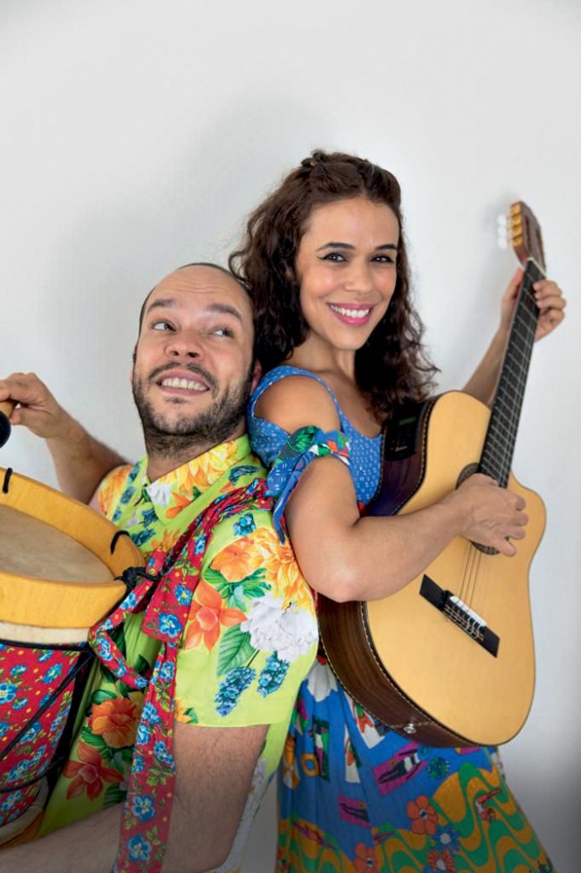 Um casal posa para a foto com roupas coloridas. A mulher segura um violão e o homem um tambor