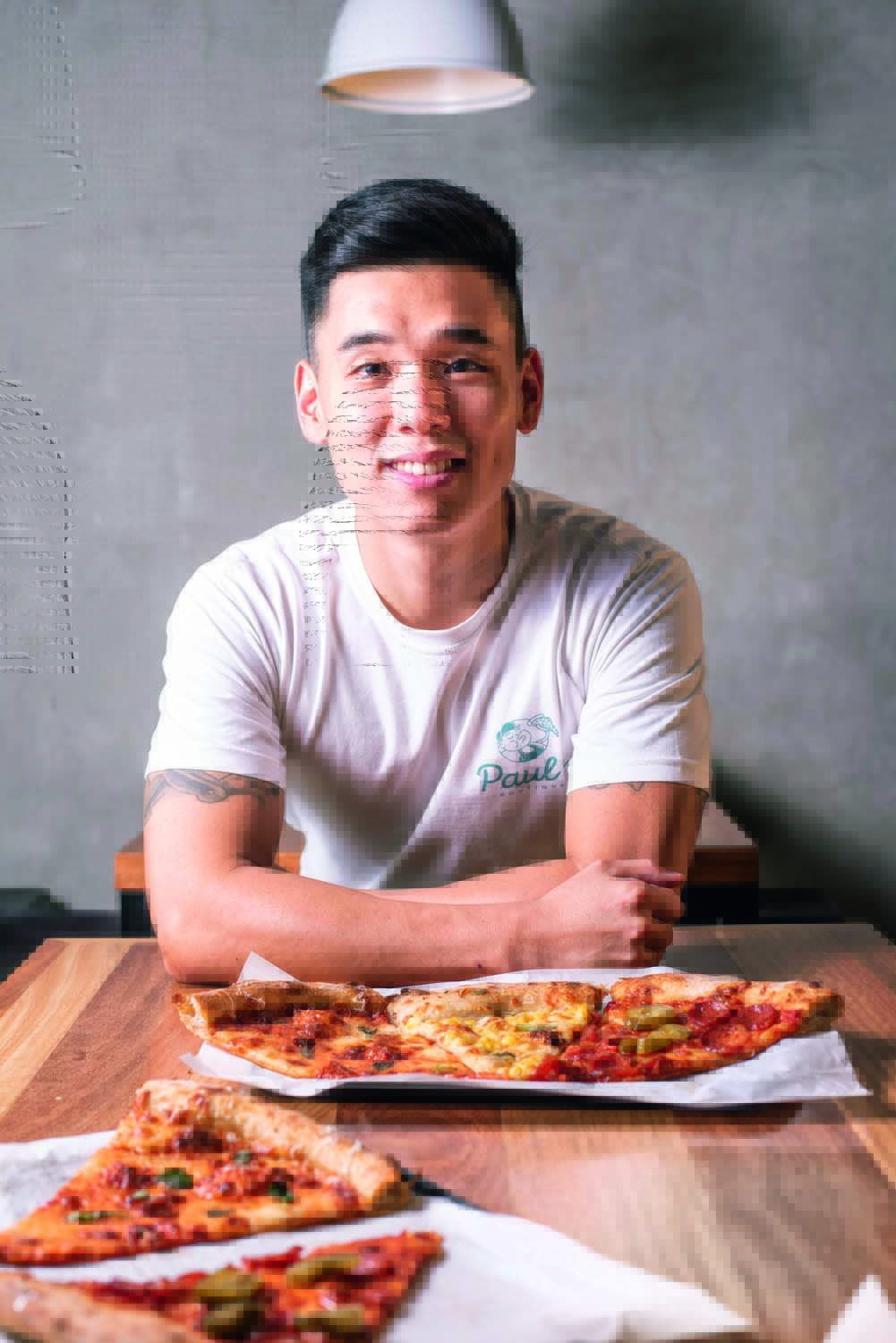 Paul Cho posa de braços cruzados sobre mesa onde estão dispostos diferentes pedaços de pizza