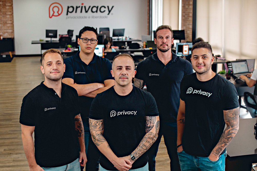 Cinco homens vestem camiseta preta com o nome "Privacy" e posam para foto em um amplo escritório. Todos são brancos e alguns têm tatuagens nos braços.
