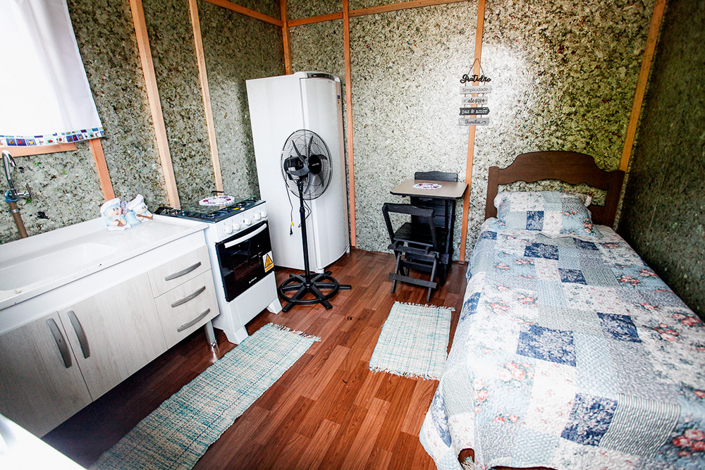 Interior de uma moradia provisória destinada à população de rua. Dentro do cômodo há uma cama de solteiro, uma geladeira, um ventilador de chão, um fogão, um armário e outros móveis. Piso é de madeira.