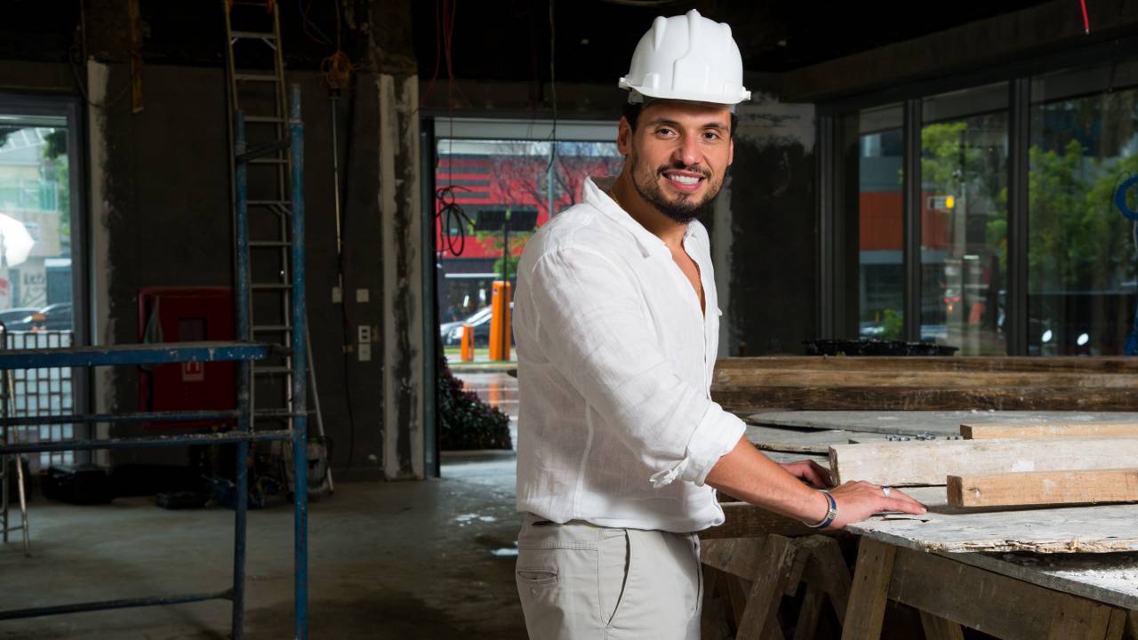 Bruno Sindona posa vestido de branco, de pé em sala ainda em obras, ao lado de uma mesa. Sorri e veste capacete de proteção, também branco.