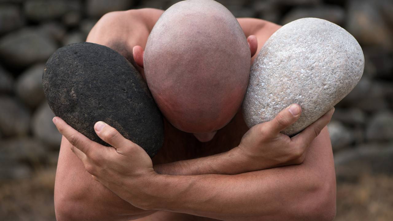 Artista Rodrigo Braga com a cabeça abaixada e abraçado com duas pedras, uma preta e uma obra. Trata-se de uma fotografia da série Ponto Zero