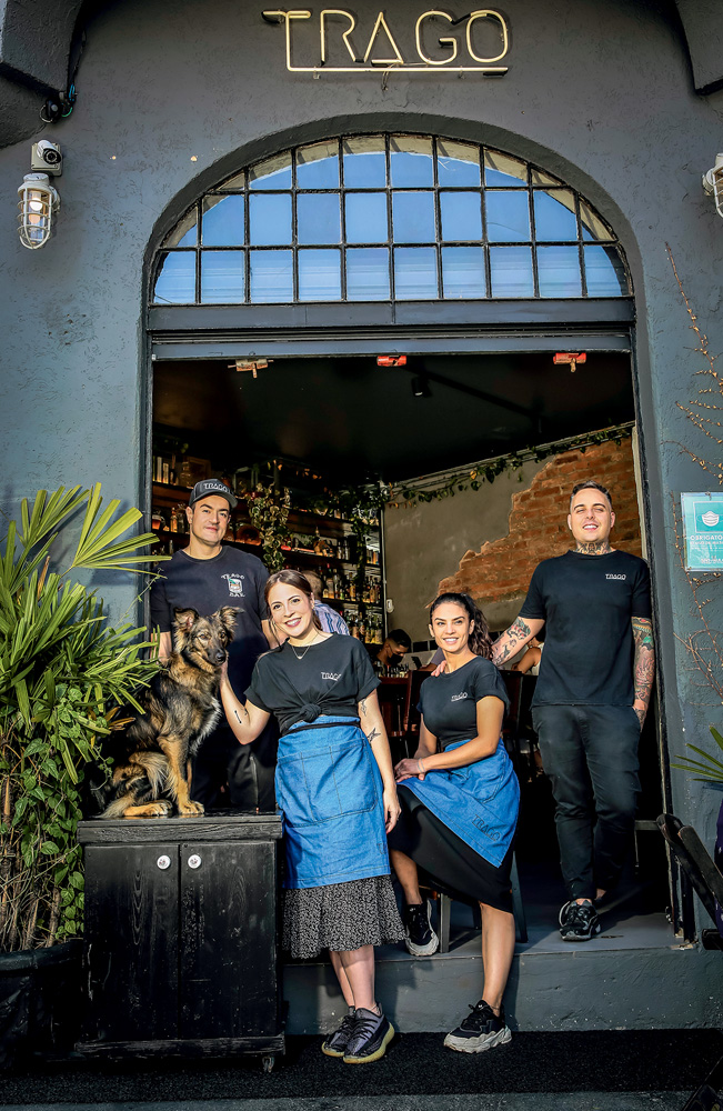 Foto mostra dois casais (os sócios do bar e suas companheiras) e um cachorro. Eles usam uniforme preto e posam na frente do Trago Bar. Todos são brancos, magros e de cabelo escuro.