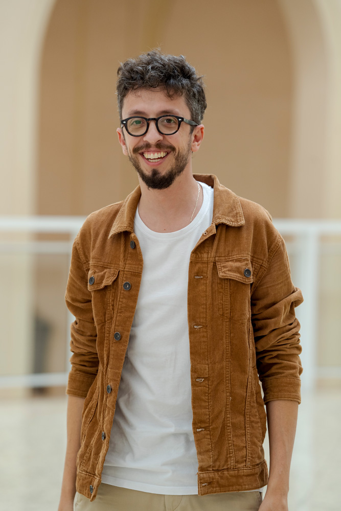 Imagem mostra homem de jaqueta bege e camiseta branca, sorrindo para a câmera.