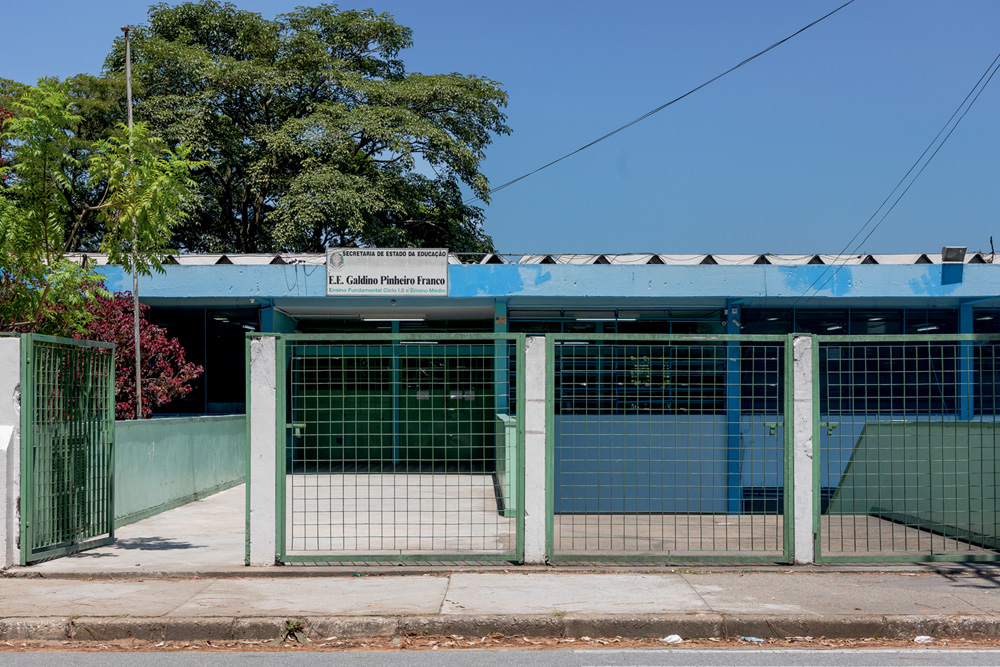 Imagem mostra fachada de escola com muro azul claro. Acima, um céu azul e uma árvore alta.