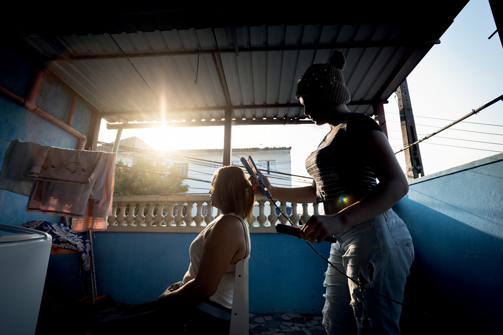 Imagem mostra mulher em pé cuidando de cabelo de outra mulher, sentada. O rosto das duas está na sombra de um raio de luz que entra diretamente na câmera, à esquerda delas.