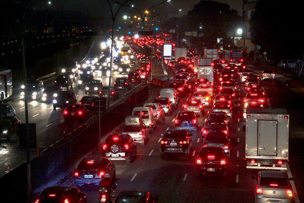 Imagem mostra estrada com aglomeração de carros. O trânsito está nos dois sentidos - a foto é noturna, portanto a iluminação é das luzes traseiras, vermelhas, da pista à direita e das luzes fronteiras, brancas, à esquerda.